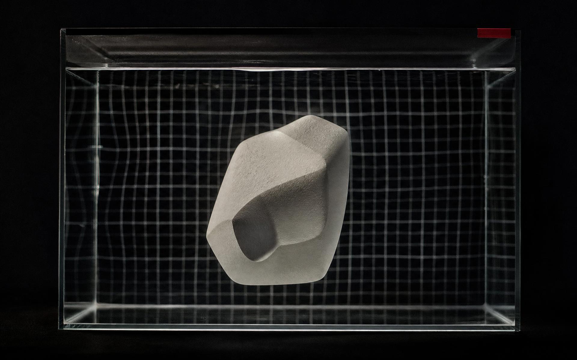 A bone-white cast block suspended in a clear water tank, bending the grid printed behind it