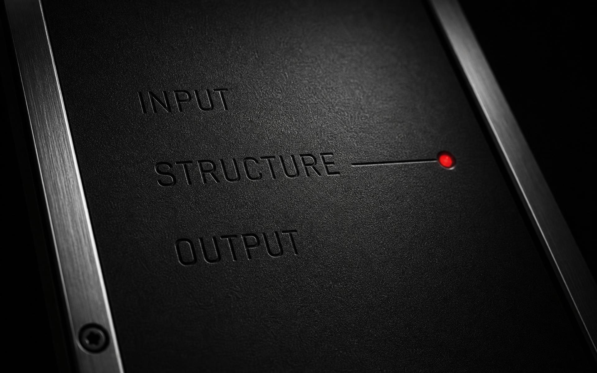 Macro of a matte black machined panel etched INPUT, STRUCTURE, OUTPUT with an oxide-red dot